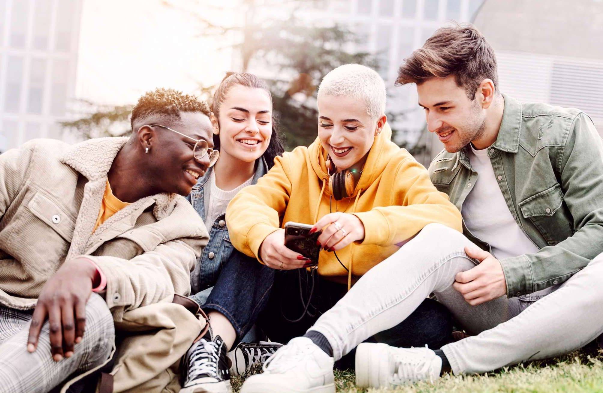 Jugendliche Frauen und Männer auf einer Wiese mit einem Handy in der Hand. Foto: adobestock Jugendliche Frauen und Männer auf einer Wiese mit einem Handy in der Hand. Foto: adobestock