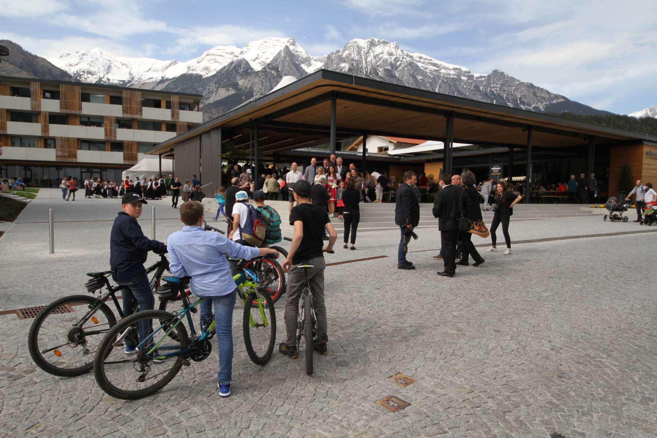 Gebäude vor Bergkulisse und davor Menschen mit Fahrrädern Gebäude vor Bergkulisse und davor Menschen mit Fahrrädern
