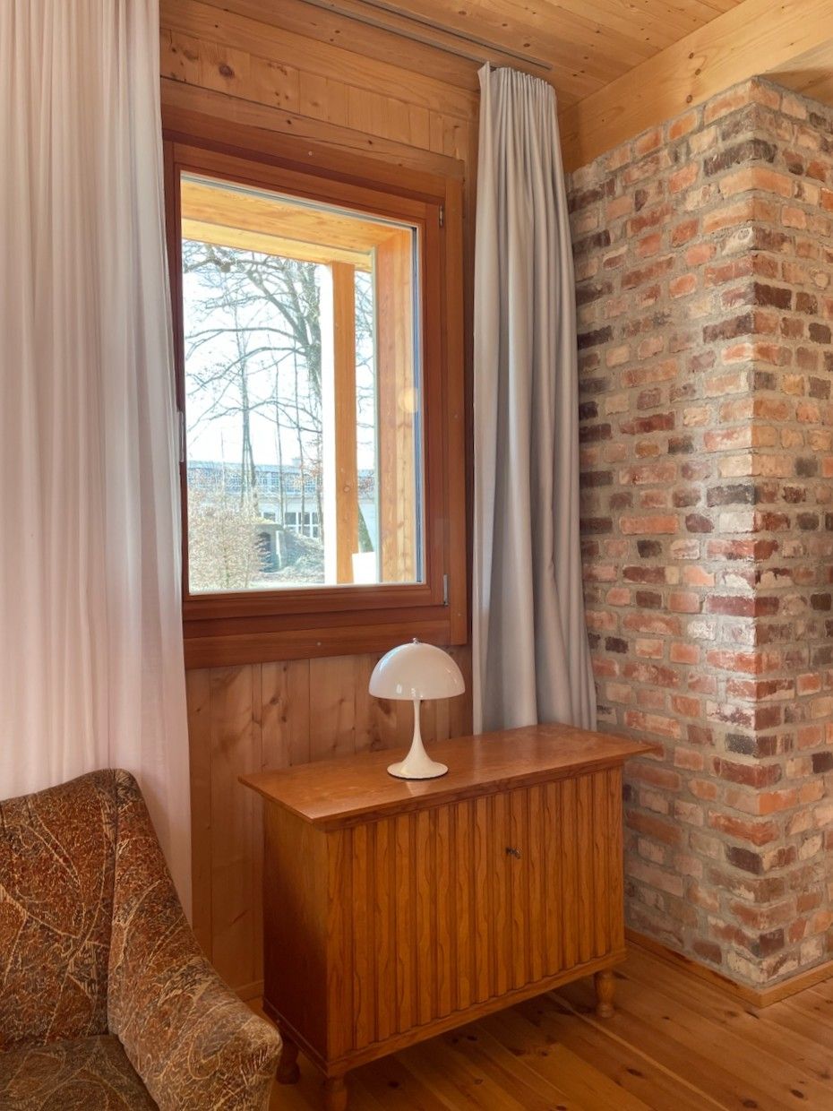 Sessel und kleine Kommode vor Fenster, Wand z.T. aus Holz und z.T. aus unverputzem Mauerwerk