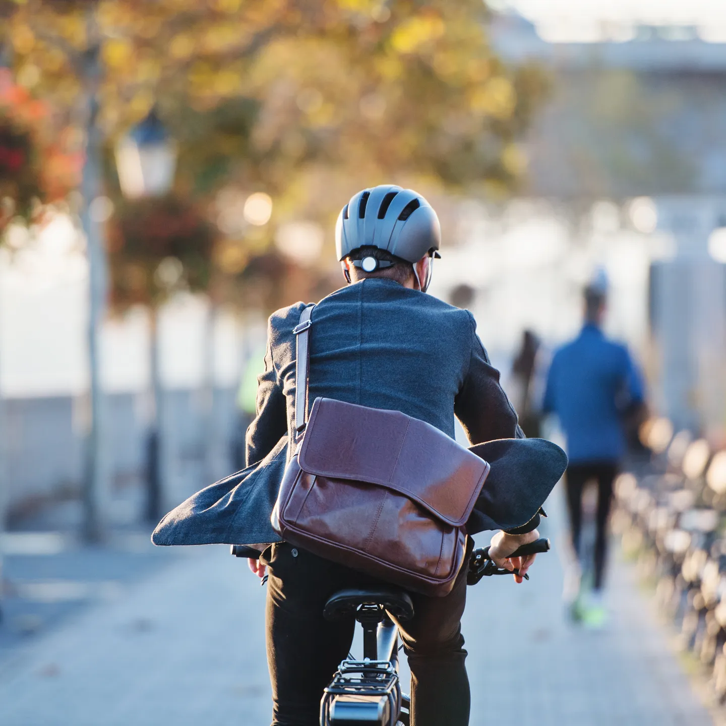 A man wearing a helmet, a suit, and carrying a briefcase over his shoulder is riding a bicycle