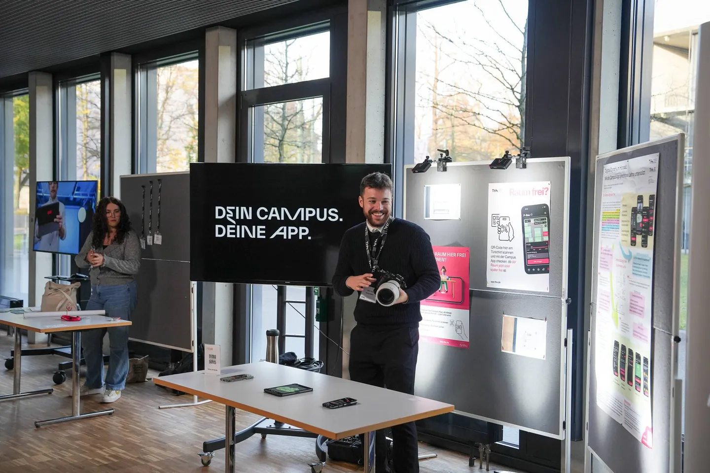 Ein Mitarbeiter des DMZ steht am App-Stand vor einem Bildschirm mit der Aufschrift „Dein Campus. Deine App.“