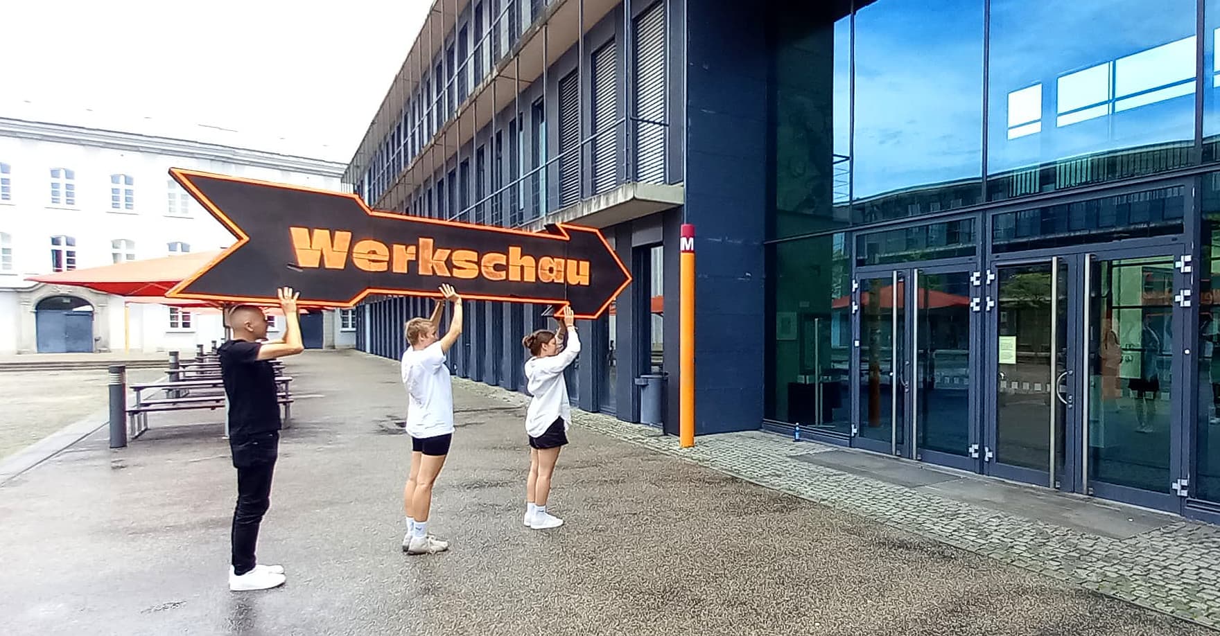 Drei Studierende im KLM-Innenhof heben einen großen Pfeil hoch mit der Aufschrift Werkschau.