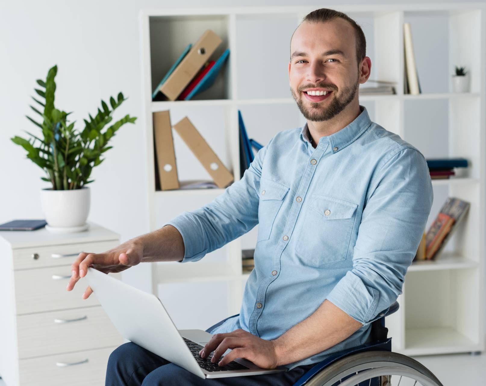 Junger Mann im Rollstuhl mit Laptop auf dem Schoß und Regal im Hintergrund