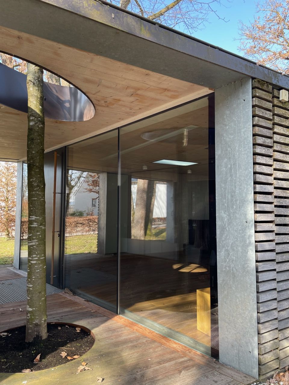 einstöckiges Gebäude mit Glasfront und Vordach mit keisförmigem Loch, durch das ein Baum wächst.